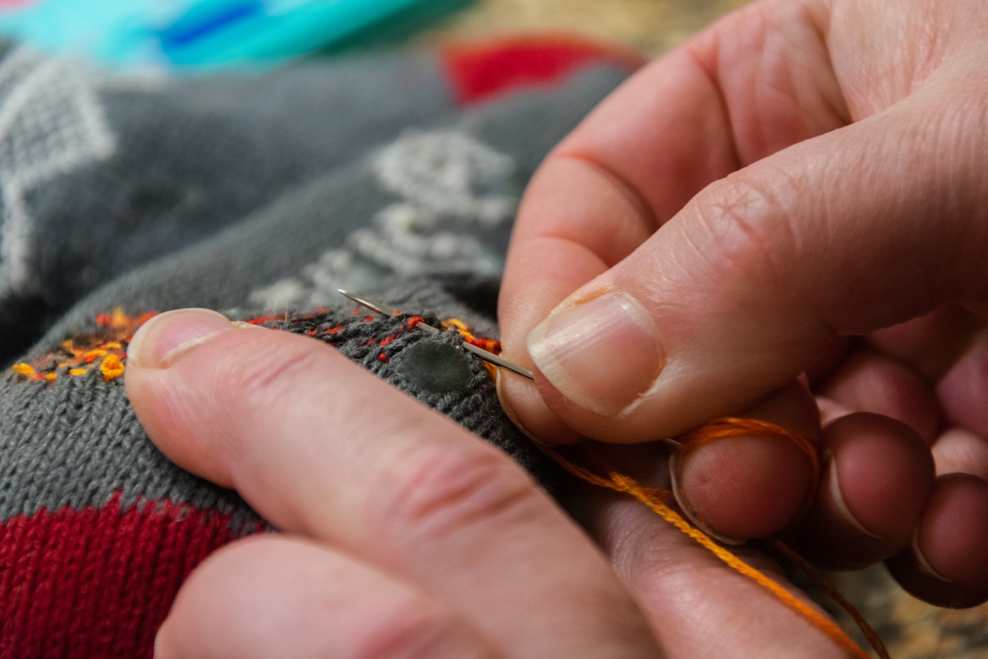 Hands sew an orange thread onto a grey knitted garment.